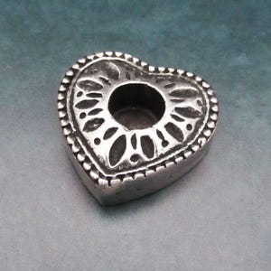 Könnte beinhalten: Ein silberner herzförmiger Kerzenhalter mit einem komplizierten Design am Rand und einer runden Öffnung in der Mitte für ein Teelicht.