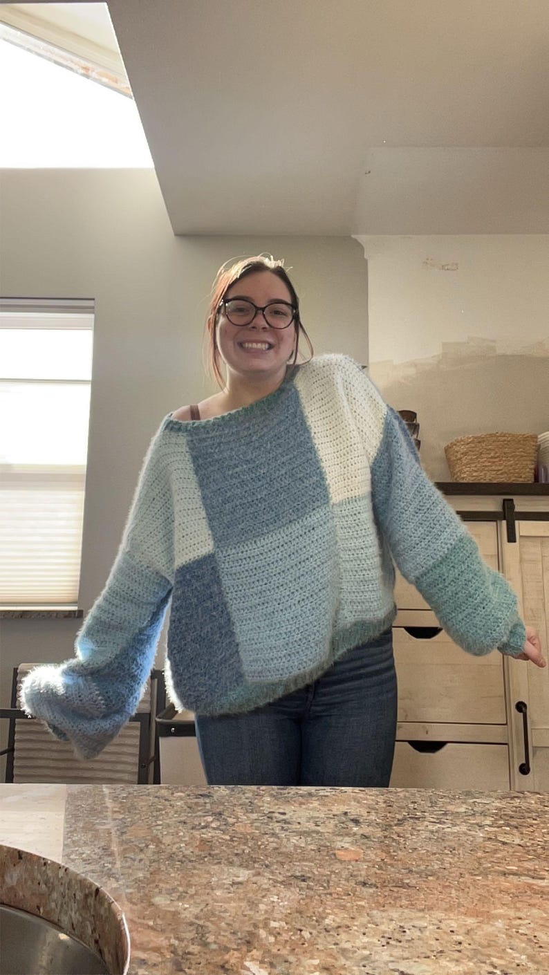 Puede incluir: Un su&eacute;ter de crochet hecho a mano, de gran tama&ntilde;o, en tonos azules y blancos. El su&eacute;ter presenta un dise&ntilde;o de patchwork con diferentes texturas y tonos de azul, de claro a oscuro. Las mangas son largas y ligeramente acampanadas.