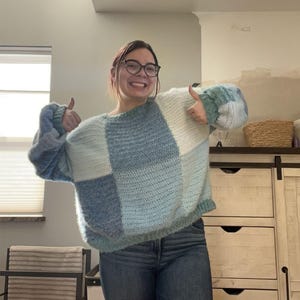 Puede incluir: Un su&eacute;ter de crochet patchwork en tonos azules y blancos. El su&eacute;ter tiene un ajuste relajado con mangas largas. El dise&ntilde;o presenta grandes bloques cuadrados de diferentes texturas y tonos, creando un look acogedor y elegante. La modelo lleva vaqueros azules.