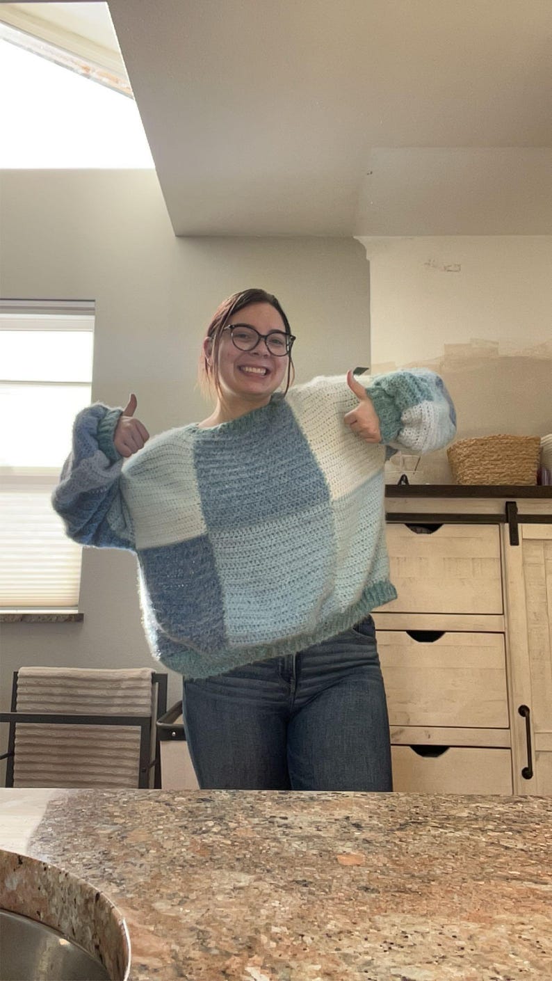 Puede incluir: Un su&eacute;ter de crochet patchwork en tonos azules y blancos. El su&eacute;ter tiene un ajuste relajado con mangas largas. El dise&ntilde;o presenta grandes bloques cuadrados de diferentes texturas y tonos, creando un look acogedor y elegante. La modelo lleva vaqueros azules.