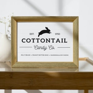 Könnte beinhalten: Ein goldfarbener Rahmen mit einem weißen Hintergrund und schwarzem Text. Das Schild lautet "EST. 1950 COTTONTAIL Candy Co." mit einer schwarzen Silhouette eines springenden Kaninchens über dem Text. Unter dem Text steht "JELLY BEANS • PEANUT BUTTER EGGS • MARSHMALLOW CHICKS".