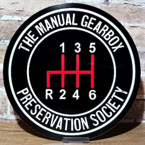 May include: Black and white round sign with the text "The Manual Gearbox Preservation Society" and a red stick shift pattern with the numbers 1, 2, 3, 4, 5, and 6.