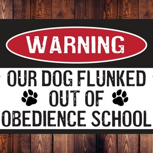 May include: A black and white metal sign with red text that reads "WARNING" in a red oval. The sign says "OUR DOG FLUNKED OUT OF OBEDIENCE SCHOOL" with two paw prints on either side of the word "OUT OF".