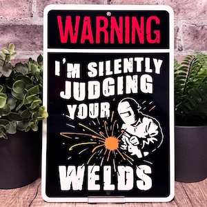 May include: Black and white metal sign with the text "WARNING I'M SILENTLY JUDGING YOUR WELDS". The sign features an illustration of a welder and sparks. The word "WARNING" is in red.