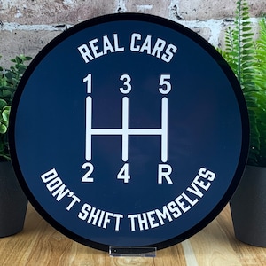 May include: A round, black and white sign with the text "REAL CARS DON'T SHIFT THEMSELVES" and a white gear shift pattern with the numbers 1, 2, 3, 4, 5, and R.