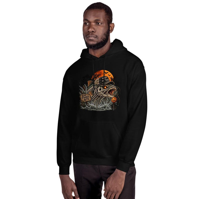 K&ouml;nnte beinhalten: Ein schwarzer Kapuzenpullover mit einem Grafikdruck eines skelettierten Fisches mit Kappe, leuchtenden Augen, einem K&uuml;rbis und einem Schild mit der Aufschrift "BAD THINGS BITE" vor einem gro&szlig;en orangefarbenen Mond und dunklem Wald.
