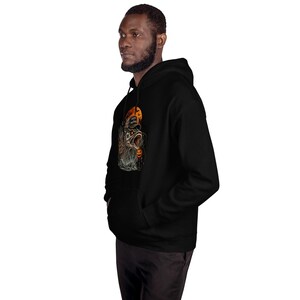 K&ouml;nnte beinhalten: Ein schwarzer Kapuzenpullover mit einem Grafikdruck eines Fisches, der eine Baseballkappe mit dem Text "HOOKED HALLOWEEN" vor einem orangefarbenen Mond tr&auml;gt, erg&auml;nzt durch einen kleinen K&uuml;rbis und Flederm&auml;use im Detail.