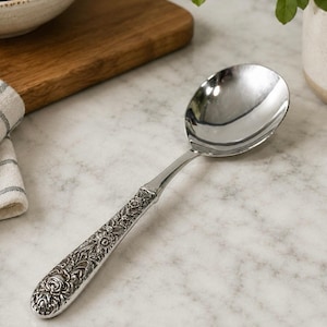 May include: A silver-toned serving spoon featuring a highly polished, smooth, rounded bowl and a textured handle decorated with an intricate, raised floral and leaf pattern that extends from the base to the neck of the utensil.