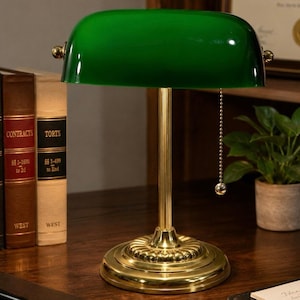May include: A banker's lamp featuring a polished brass base and a cylindrical stem with a fluted design. The lamp has a curved, emerald green glass shade and a gold-toned metal pull chain with a small spherical end for turning the light on and off.