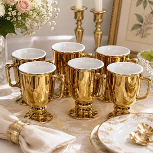 May include: A set of six metallic gold mugs with white interiors, each featuring a pedestal base and a side handle. The mugs are arranged in a group, showcasing a reflective, polished finish that catches the light, creating a bright and uniform appearance.