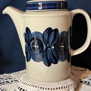 May include: A cream-colored ceramic teapot featuring a cylindrical body and a matching lid. The teapot is decorated with a horizontal band of dark blue, hand-painted floral motifs. It includes a curved handle and a pointed pouring spout.