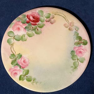 May include: Round, hand-painted ceramic plate with a floral design. The plate features pink and red roses with green leaves arranged around the edge. The background has a gradient of yellow, pink, and green hues.