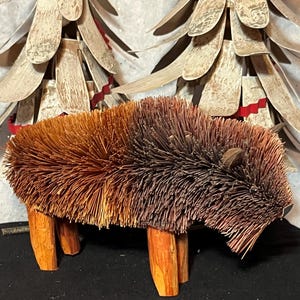 May include: A handcrafted bison figurine constructed from natural brown pine needles and twigs. The body features a dense, textured coat of dark and light brown needles, while the four legs are made from smooth, light-colored wooden branches.