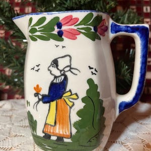 May include: A white ceramic pitcher with a blue handle and rim. The pitcher is decorated with a folk art design featuring a woman in traditional clothing, flowers, and birds. The handle and rim are painted blue.
