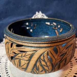 May include: A handcrafted ceramic bowl with a leaf design. The bowl features a brown exterior with cut-out leaf patterns and a deep blue interior. The rim is a darker brown, and the bowl is sitting on a white lace doily.