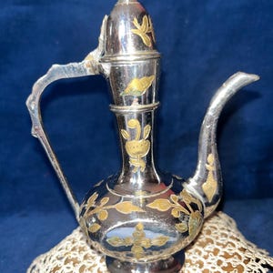 May include: A silver-toned metal ewer featuring a tall, slender neck and a curved handle. The surface is decorated with engraved gold-colored floral and leaf patterns. The vessel has a pointed spout and a small, finial-topped lid at the peak.