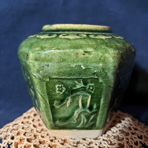 May include: A hexagonal ceramic jar with a glossy, deep green crackle glaze finish. The exterior features embossed floral motifs on each side panel. The rim is circular, and the base is unglazed, showing a light beige color. The surface has visible fine lines throughout the glaze.