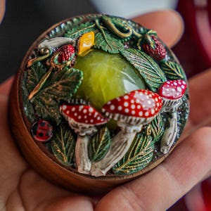 May include: A round, wooden trinket box with a detailed, nature-themed design. The lid features a central green stone surrounded by sculpted mushrooms, leaves, and berries. The box is held in a hand, with a blurred background.