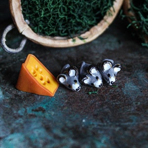 May include: Three small gray ceramic mice figurines with black eyes and noses, lined up on a dark green plate with a wedge of orange cheese.