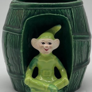 May include: A green ceramic planter shaped like a barrel with a green elf figurine sitting inside. The elf is wearing a green hat and green clothing.
