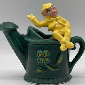 May include: A green ceramic watering can with a yellow elf sitting on top. The watering can has a flower design and a spout. The elf is wearing a yellow hat and has a mischievous expression.