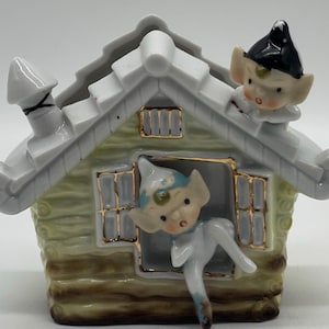 Puede incluir: Una casa de cerámica blanca con un diseño de cabaña de troncos verde y dorado. Dos elfos asoman por las ventanas. Un elfo está en el techo.