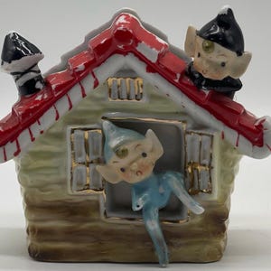 May include: A ceramic figurine of a small house with a red roof and white trim. Two elves are peeking out of the windows. One elf is wearing a blue suit and the other is wearing a black suit.