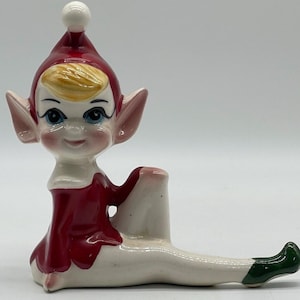 May include: A ceramic Christmas elf figurine with a red hat, white trim, and a white pom-pom. The elf is sitting with its legs stretched out and wearing a red dress with white trim and green shoes.