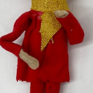 May include: A vintage felt elf doll with a red outfit, gold trim, and a smiling face. The elf has orange hair and is wearing a gold scarf.