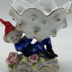 May include: A porcelain figurine of a gnome holding a white shell-shaped dish. The gnome is wearing a red hat, blue and white patterned clothing, and black boots. The dish has a gold rim and is decorated with gold dots. The figurine is sitting on a base with pink roses and green leaves.