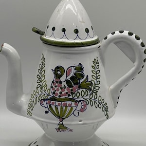 May include: White ceramic teapot with a green and pink bird design. The teapot has a lid with a knob in the shape of a clover. The teapot has a spout and a handle.