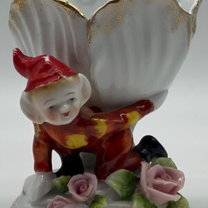 May include: A porcelain figurine of a red and yellow elf holding a white shell-shaped vase with gold trim. The elf is kneeling on a base with pink roses and green leaves.