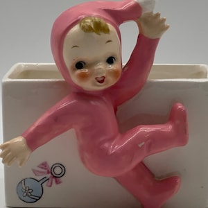 May include: A pink ceramic planter shaped like a baby in a pink onesie. The baby is holding its head up and has a white cap on. The planter has a white background with a blue and pink rattle design.