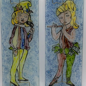 May include: Two vintage glass panels with painted images of figures. The panel on the left features a jester with a yellow hat and purple and blue clothing. The panel on the right features a figure with blonde hair playing a flute. Both panels have a blue background with white bubbles.