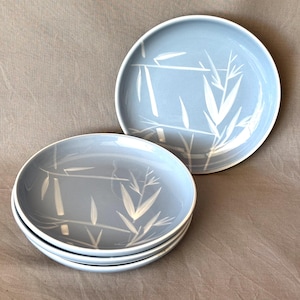 May include: A set of four light blue ceramic plates with a white bamboo design. One plate is separate, and the others are stacked. The plates have a simple, elegant design, perfect for serving food.
