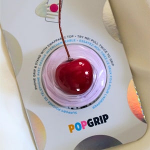 May include: A red cherry PopGrip phone grip with a long stem, presented on its original packaging. The packaging features the words "PopGrip" and "Phone Grip & Stand".