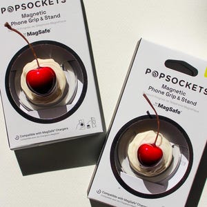 Peut inclure: Deux boîtes PopSockets avec un motif de cerise. Chaque boîte présente un fond blanc avec le logo PopSockets et des informations sur le produit. Le motif de la cerise est une cerise rouge avec une tige marron sur une base blanche.