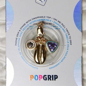 May include: A gold PopGrip phone stand with a seashell charm, a blue and purple gem, and a white gem. The PopGrip is on a white background with the text "PopSockets" and "PopGrip" printed on it.