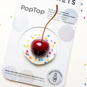 Peut inclure: Un support et support de téléphone PopSockets PopTop avec un motif de cerise. La cerise est rouge brillant avec une tige marron, posée sur une base blanche avec des sprinkles colorés. L'emballage comprend le texte "POPSOCKETS" et "PopTop".