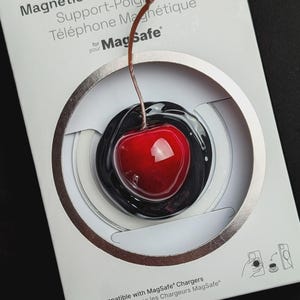Peut inclure: Un support magnétique pour téléphone PopSockets en forme de cerise rouge brillante avec une tige brune, fixé sur une carte de vente blanche. L'emballage porte le texte PopSockets Magnetic Phone Grip & Stand for MagSafe et des icônes.