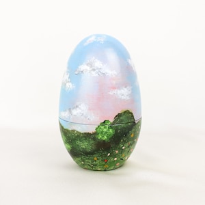 May include: A hand-painted wooden egg with a blue sky, pink clouds, green grass, and trees. The egg is a decorative item.