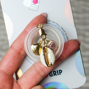 May include: A gold-colored phone grip with a white seashell charm, a pink heart charm, and a gold charm. The grip is attached to a clear plastic base. The packaging for the grip is in the background and says "POPSOCKETS" and "GRIP".