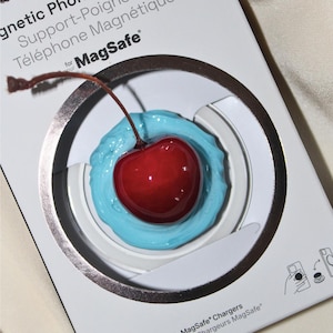 May include: A phone grip and stand with a cherry design. The red cherry is set in blue material, inside a white and silver circular frame. The packaging reads "Magnetic Phone Grip & Stand" and "MagSafe".