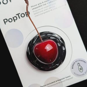 Pode incluir: Um suporte para telemóvel PopSockets PopTop com uma cereja vermelha brilhante e um caule castanho fixado numa base preta em espiral. O produto está montado numa embalagem branca com o texto POPSOCKETS, PopTop e Bonus Accessory.