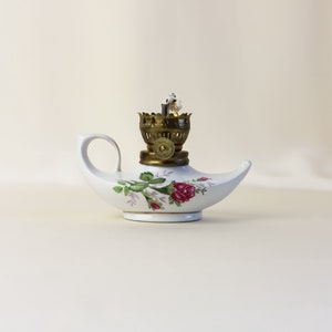 Op de afbeelding: Een witte keramische olielamp met een bloemenontwerp met roze rozen en groene bladeren. De lamp heeft een messing lont houder en een tuit om olie in te gieten.