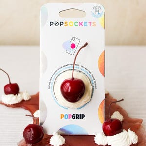 Op de afbeelding: Een PopSockets PopGrip telefoonaccessoire met een kersenthema. De PopGrip heeft een glanzend rood kersendesign op een witte basis, met een bijpassende kers op een rode, stroperige ondergrond. De verpakking is zichtbaar, met het PopSockets-logo.