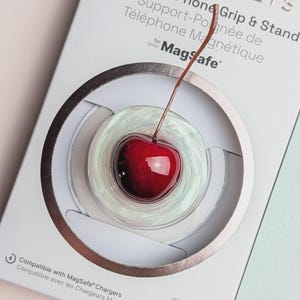 May include: A PopSockets magnetic phone grip and stand in retail packaging. The grip features a clear, iridescent base with a realistic red cherry and stem on top. The white packaging includes the text PopSockets, Magnetic Phone Grip & Stand, and MagSafe.