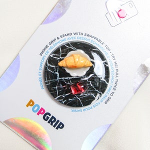 Könnte beinhalten: Ein schwarzer und weißer Marmor-Handy-Griff mit einem Croissant auf einem weißen Teller. Der Griff ist ein Pop-up-Stil und hat eine austauschbare Oberseite. Der Text auf der Verpackung lautet: "Phone Grip & Stand with Swappable Top. Try Me! Pull Twice to Grip. Poignée et Support de Téléphone avec Dessus Échangeable. Essayez-moi! Tirez Deux Fois pour Saisir."