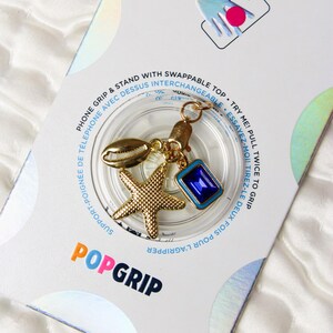 May include: A gold phone grip with a starfish, seashell, and blue gem charm. The grip is attached to a clear plastic base with the text "POPGRIP" printed on it. The packaging includes the text "PHONE GRIP & STAND WITH SWAPPABLE TOP. TRY ME! PULL TWICE TO GRIP. SUPPORT-POIGNÉE DE TELEPHONE AVEC DESSUS INTERCHANGEABLE. ESSAYEZ-MOI! TIREZ-LE DEUX FOIS POUR L'AGRIPPER."