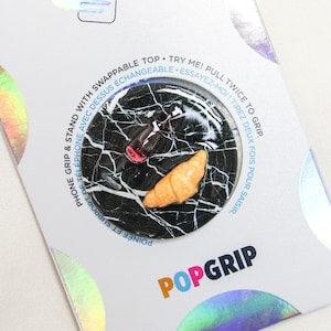 Könnte beinhalten: Ein schwarzer und weißer Marmor-Handy-Griff mit einem Croissant und einem Glas Wein. Der Griff ist rund und hat eine ausklappbare Funktion. Der Text "POPGRIP" ist auf der Verpackung aufgedruckt.